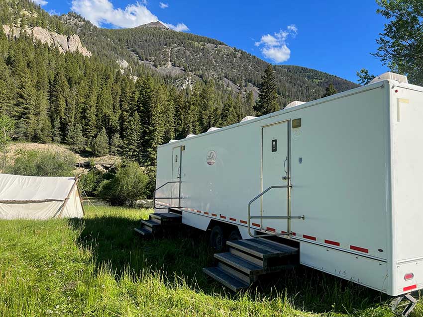 Mobile shower trailers with steps in a grassy area near a forested mountain under a blue sky with a few clouds.